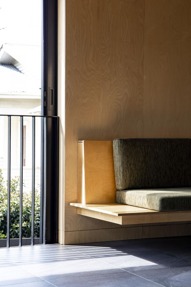 A bright, minimalist interior nook featuring a built-in timber bench seat with dark upholstered cushions against full-height timber veneer wall panelling. An open glazed doorway with black metal framing and an external balustrade brings in strong sunlight, casting linear shadows across the dark tiled floor.