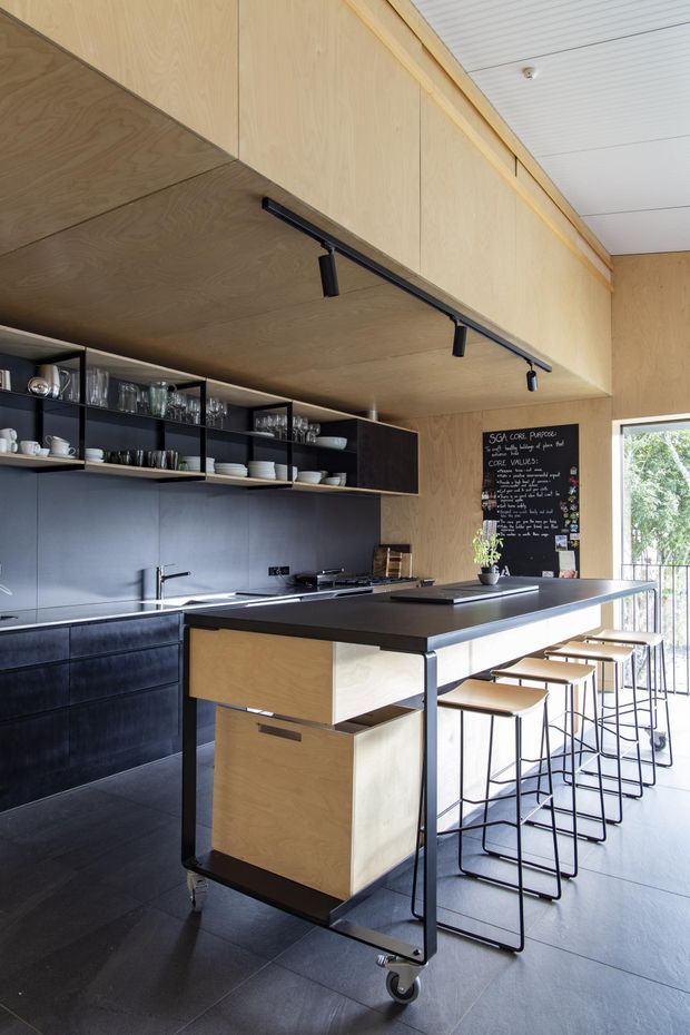 A compact contemporary kitchen with extensive light timber plywood wall and ceiling panels, dark cabinetry, open shelving, and a large black island with integrated timber storage. A mobile black-framed trolley sits in the foreground, while a row of slim bar stools lines the island beside a bright window. Track lighting and a chalkboard add practical café-like character.