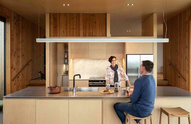 Warm, timber-lined open-plan kitchen dominated by a large central island with an integrated sink, stainless-steel benchtop, pale wood cabinetry, and island seating. A couple occupies the kitchen, with a suspended linear light above the island, built-in shelving, and a refrigerator visible in the background. The space has a compact, highly integrated contemporary fit-out with extensive vertical timber wall panelling.