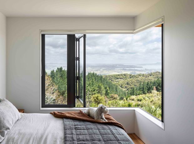 A bright, minimalist bedroom with a neatly made bed positioned beside large corner glazing. Black-framed casement windows open toward a broad elevated landscape view of trees, hills, and distant water. The room has smooth light-grey walls, a white ceiling, and warm timber flooring, with a small dog resting on the bed.
