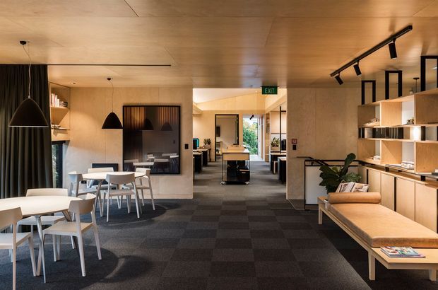 A warm, contemporary commercial interior resembling a café, hospitality venue, or shared workspace. The open-plan space features pale timber wall and ceiling lining, dark carpet tiles, light timber built-in shelving and cabinetry, white dining tables and chairs, pendant lighting, black track lighting, and a long upholstered bench seat. A central corridor leads toward glazed doors and additional work or dining areas.