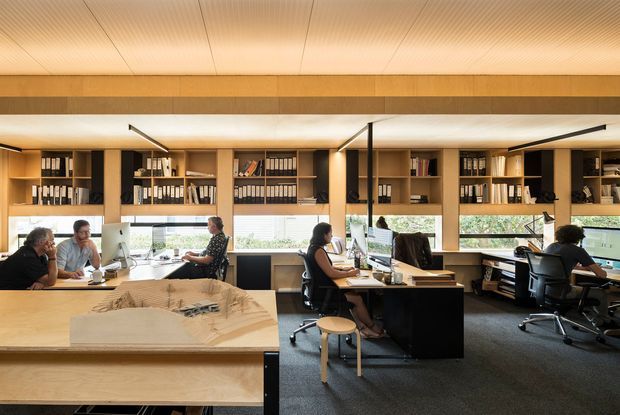 Open-plan professional office with several workstations, people working at desks, extensive built-in shelving and storage, a large architectural model on a central worktable, warm timber wall and ceiling finishes, and large windows providing daylight. The space has a refined, contemporary architectural studio character with dark carpet and black workstation elements.
