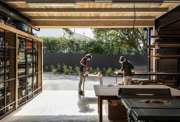 Open-plan workshop or studio with a large raised roller door opening onto a landscaped outdoor work area. The space has exposed timber ceiling beams and decking, polished concrete flooring, dark built-in shelving and cabinetry, metal-framed storage, workbenches, and large workshop equipment. Two people are working at an outdoor table, while daylight and greenery are visible beyond the opening.