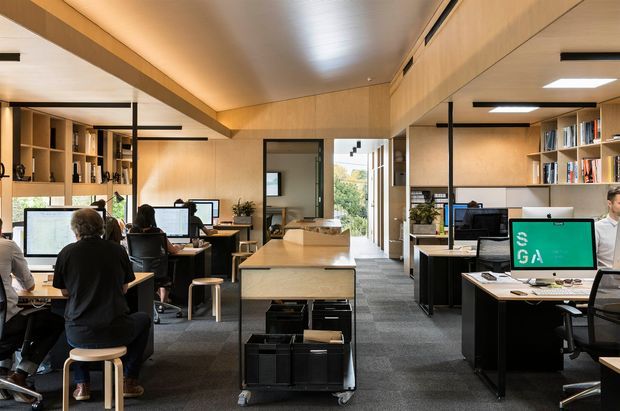 Open-plan contemporary office interior with multiple computer workstations, shared desks, shelving, and staff working throughout the space. Warm timber wall and ceiling lining contrasts with dark cabinetry, grey carpet flooring, black metal-framed glazing, and exposed linear lighting. The office has a long, organized layout with a central shared workbench and views through glazing toward greenery.