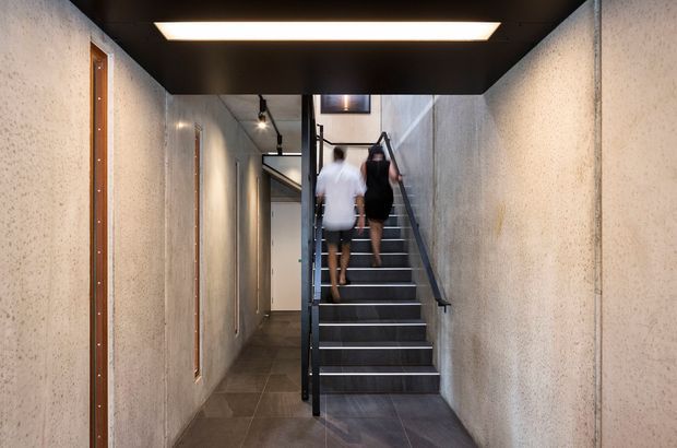 A contemporary interior stairway and hallway with dark tiled flooring, exposed textured concrete walls, black metal stair framing and balustrade, timber-framed narrow windows, and a dark ceiling with a large rectangular recessed light. The stair rises toward an upper level, with a corridor visible alongside it.