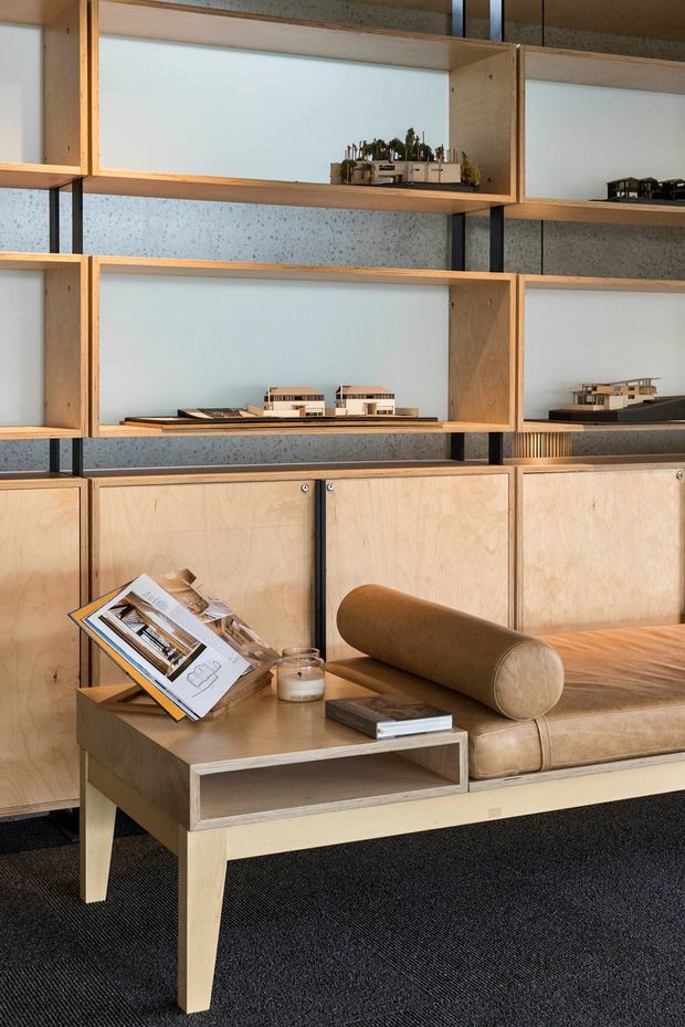 A warm, refined interior vignette featuring a built-in timber display wall with open shelving and lower storage cabinets. Architectural scale models are arranged on the shelves, suggesting a design studio or architectural showroom. In front is a tan leather built-in bench or sofa with cylindrical bolster, paired with a light timber coffee table holding architectural publications. The space has a speckled grey wall backing, dark carpet, and black vertical structural supports.