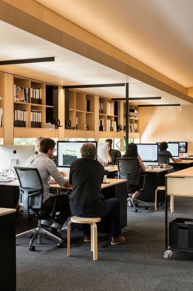 A warm, contemporary open-plan office with several workstations, desktop monitors, ergonomic office chairs, built-in timber shelving filled with binders, and integrated storage. The room features dark carpet flooring, pale timber cabinetry and wall cladding, exposed black structural beams, and a ribbed timber ceiling with linear pendant lighting. Several people are working at desks, creating an active professional workspace.