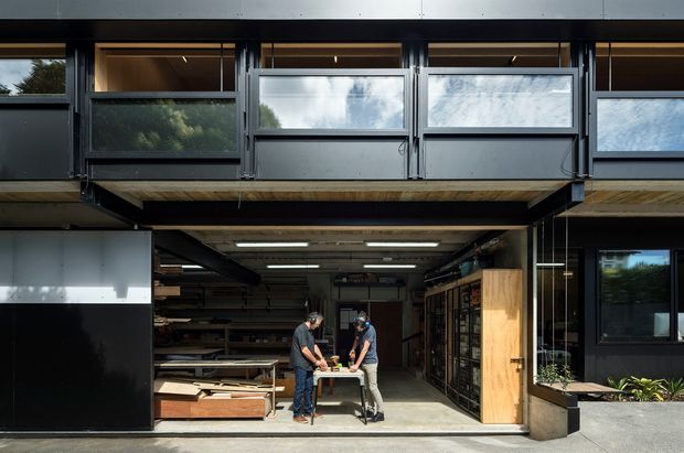Exterior view of a contemporary multi-level workshop or studio building with a large open ground-floor work area, clerestory-style windows, dark metal framing, and timber soffit detailing. Two people are working at a central table inside; shelving, timber materials, and workshop equipment are visible. The upper level projects over the open workshop frontage, creating a covered entry or loading zone.