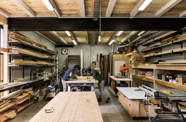 A spacious woodworking workshop or fabrication studio filled with timber sheets, boards, shelving, workbenches, and stationary woodworking machinery. Two people are working at a central bench. The space has exposed timber ceiling joists and decking, dark structural beams, concrete flooring, industrial fluorescent lighting, and extensive open storage.