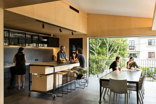 Open-plan contemporary kitchen and dining area with warm timber cabinetry, dark benchtops, integrated open shelving, and a mobile kitchen island with bar seating. A large glazed opening connects the dining zone to a balcony or terrace, bringing in abundant daylight and views of greenery and neighbouring buildings. Track lighting and streamlined detailing create a refined, functional interior.