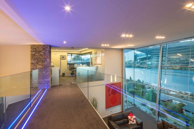 A contemporary multi-level interior overlooking a waterfront, with a carpeted mezzanine or landing, glass balustrades, extensive floor-to-ceiling glazing, exposed stacked-stone feature columns, recessed ceiling lights, and blue integrated accent lighting. A lounge area is visible below, while the view through the glazing shows water and urban buildings.