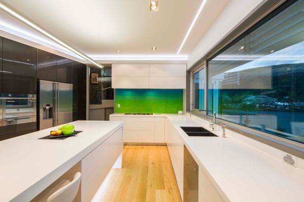 Contemporary galley-style kitchen with a large white island, white perimeter cabinetry, dark glossy cabinetry and integrated stainless-steel appliances. Full-height glazing along one side overlooks a wooded waterfront or hillside setting. Linear LED lighting and light timber flooring create a bright, refined interior.