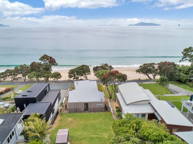 Elevated aerial view across several residential properties toward a sandy beach and calm ocean, with distant islands on the horizon. The homes have contemporary low-pitched metal roofs, landscaped gardens, lawns, and a coastal suburban setting under a partly cloudy sky.