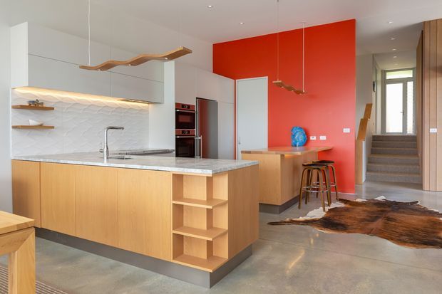 Bright contemporary open-plan kitchen with a large light-wood island, pale stone benchtop, white textured tile splashback, integrated appliances and open shelving. A vivid red feature wall, sculptural pendant lights, polished concrete flooring and a compact dining or breakfast area add visual contrast. The kitchen opens toward a stairway and timber-lined entry zone.
