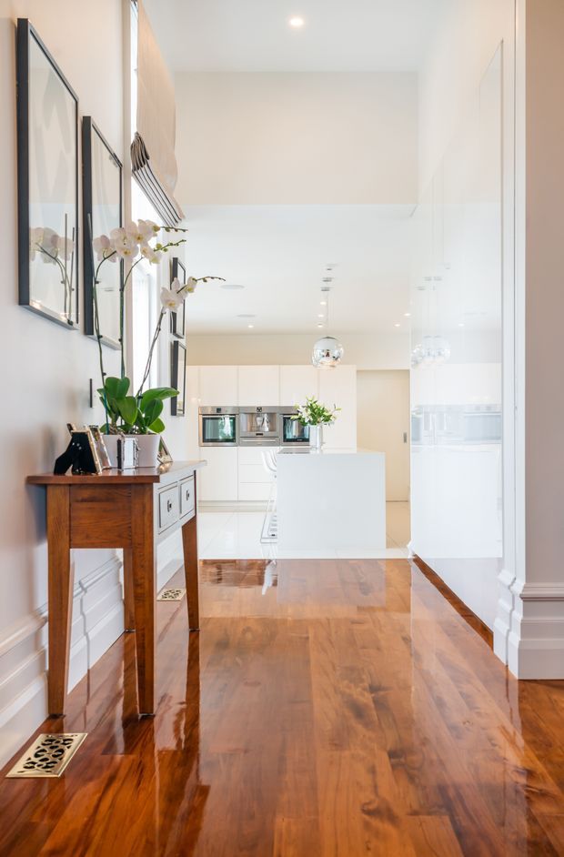 Bright, polished residential interior viewed from an entry hall toward an open white kitchen. The space features glossy warm timber flooring, white walls and cabinetry, a central island, integrated appliances, pendant lighting, framed wall art, and a traditional wooden console table with orchids. A large reflective glass or mirror panel on the right visually expands the hallway.
