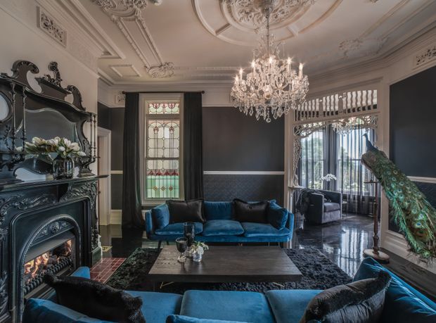 A richly detailed formal living room with dark walls, ornate plaster ceiling mouldings, a large crystal chandelier, decorative stained-glass window, carved black fireplace, and glossy dark flooring. Deep teal velvet sofas and armchairs surround a central coffee table, while a peacock display stands near the tall glazed doors. The room has an opulent, historic character with dramatic contrast and highly decorative detailing.