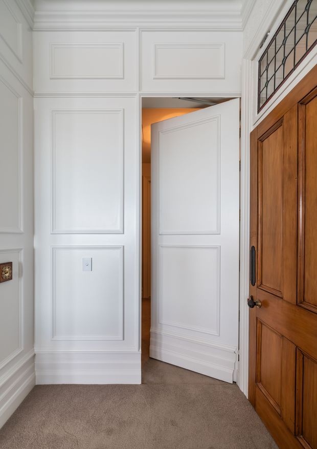 A refined traditional interior hallway or landing with white raised-panel wall panelling, decorative mouldings, a coffered or layered cornice ceiling, and beige carpet. A white panelled door stands open to a warmly lit adjoining space, while a substantial dark timber panelled door and leadlight-style glazing add contrast on the right.