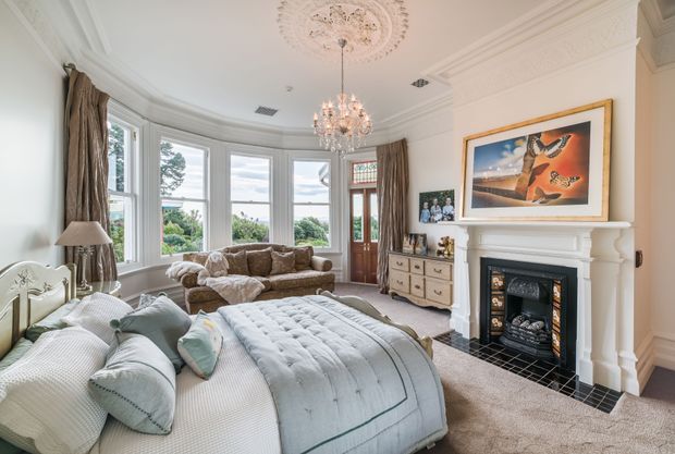 A bright, elegant bedroom in a traditional period-style home, featuring a large upholstered bed, bay-window seating area, ornate ceiling medallion, crystal chandelier, decorative cornices, layered curtains, a classic white fireplace surround, and framed artwork. The room has a soft neutral palette with pale blue-green bedding and plush beige carpet.