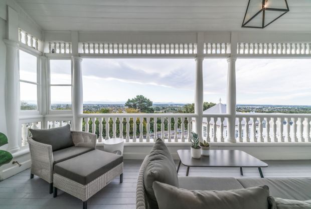 Bright enclosed veranda or sunroom with white-painted timber columns, balustrades, louvred detailing, and a white paneled ceiling. Wicker lounge seating with grey cushions faces expansive views through large windows and open balcony sections. The space has a relaxed coastal character and abundant natural light.