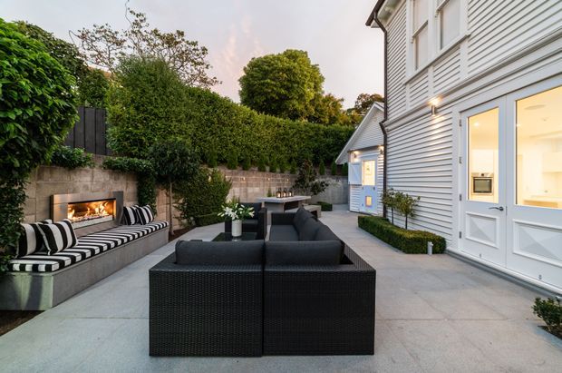 A landscaped outdoor entertaining courtyard beside a light-grey weatherboard house. The paved terrace features black woven outdoor sofas, a striped built-in bench, a wall-mounted fireplace with a lit fire, a compact outdoor bar or serving counter, clipped hedges, and concrete retaining walls. The house has white-trimmed windows and doors, exterior wall lighting, and a small gabled outbuilding or extension in the background.