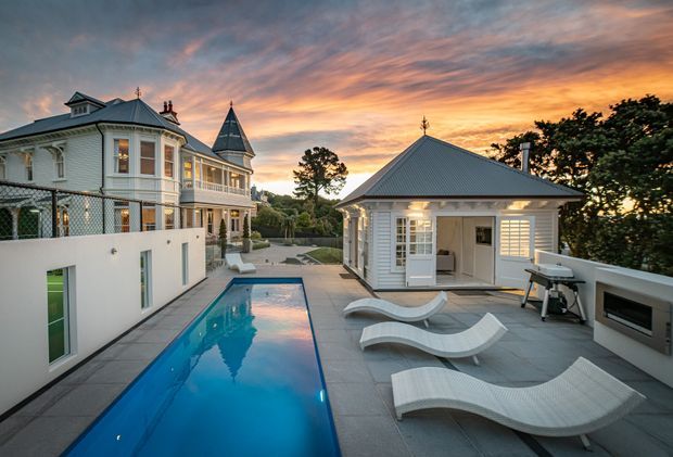 A refined traditional-style residential property photographed at sunset, featuring a narrow rectangular swimming pool, sculptural white loungers, and a paved terrace. The main house has ornate pale cladding, bay windows, balconies, decorative trim, and a steep turret roof, while a separate pool pavilion with a hipped metal roof and illuminated windows overlooks the pool.