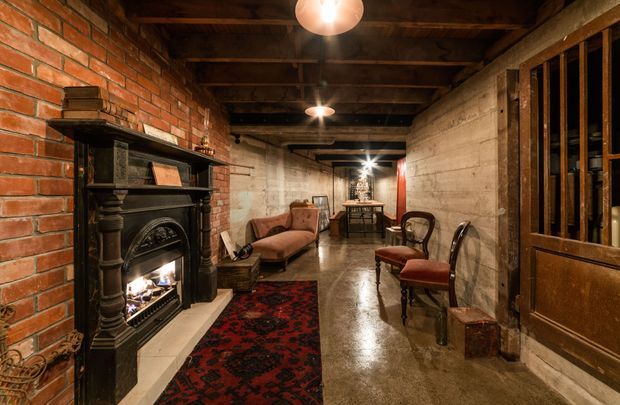 A long, narrow basement-style interior with exposed red brick and raw concrete walls, dark exposed timber ceiling beams, and a polished concrete floor. The space is furnished as a warm lounge and entertaining area with a black ornate fireplace, vintage seating, a red patterned rug, a small dining table at the far end, and dark timber-framed glazing or doors. Pendant and wall lighting create an intimate, atmospheric setting.