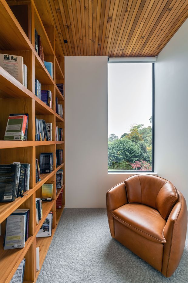 A compact reading nook or home library with floor-to-ceiling timber bookshelves filled with books, a warm timber slatted ceiling, pale walls, grey carpet, and a cognac leather lounge chair beside a tall window overlooking greenery. The space has a warm, quiet, study-like atmosphere.