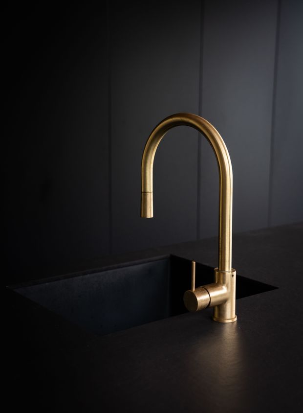 A close-up product-style image of a minimalist arched kitchen mixer tap in a warm brushed brass finish, mounted on a dark countertop beside a deep black undermount sink. The background features dark vertical wall panelling, creating a dramatic, refined and moody composition.