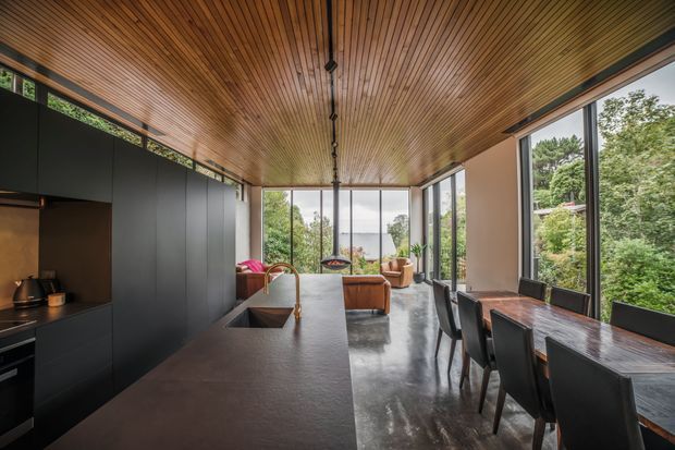 A contemporary open-plan kitchen, dining, and living space with a long dark island and matte black cabinetry, warm timber slatted ceiling, polished concrete floor, extensive floor-to-ceiling glazing, and a dark timber dining table. Lush greenery and a glimpse of water are visible beyond the windows.