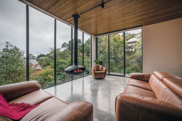 A contemporary living room with full-height glazed walls overlooking a leafy suburban setting. The room features a polished concrete floor, warm timber slatted ceiling, a suspended black fireplace, and large brown leather sofas with a matching armchair. The open corner glazing and restrained material palette create a spacious, warm, indoor-outdoor atmosphere.