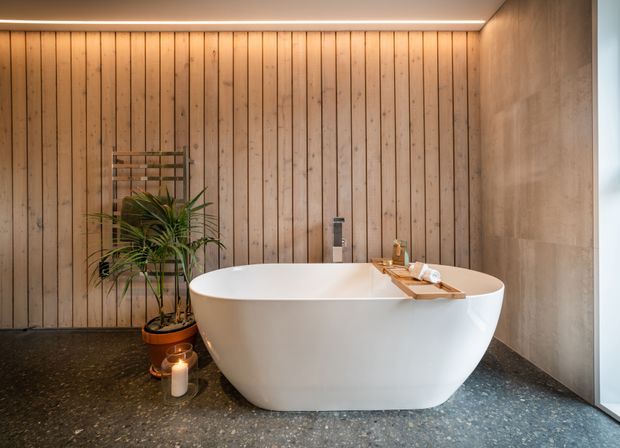 A contemporary bathroom featuring a freestanding white oval bathtub, timber vertical wall panelling, a dark speckled stone or terrazzo floor, and a smooth grey concrete-look wall. Warm concealed ceiling lighting, a timber bath tray, indoor plant, and candles create a calm spa-like atmosphere.