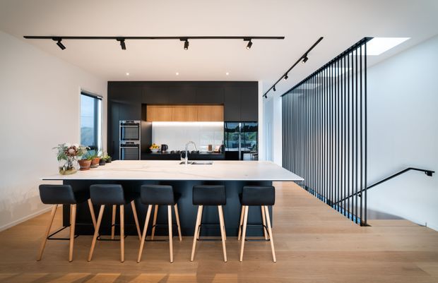A bright contemporary kitchen and dining space with a long white island and six black bar stools, light timber flooring, and dark integrated cabinetry. Black track lighting runs across the ceiling, while a timber feature panel warms the kitchen joinery. A dramatic black vertical balustrade separates the kitchen from a stairwell, with a skylight adding natural light.