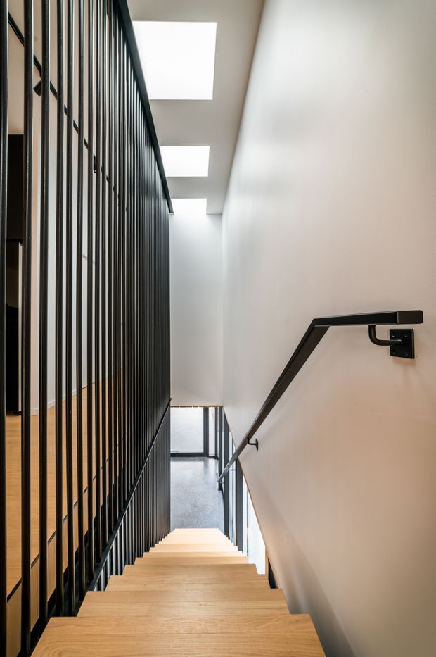 A contemporary stairway viewed from the upper level, with light timber treads, a black metal vertical-rod balustrade, and a matching angular wall-mounted handrail. White walls and several rectangular skylights create a bright, minimalist corridor-like stairwell, with a lower landing visible below.