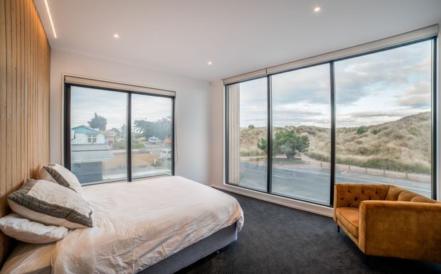 A contemporary bedroom with a large bed, dark carpet, warm vertical timber wall panelling, and expansive floor-to-ceiling glazing. A mustard velvet armchair sits beside the windows, which overlook a coastal dune landscape and neighbouring houses. Recessed ceiling lights and slim black window frames create a clean, modern interior.
