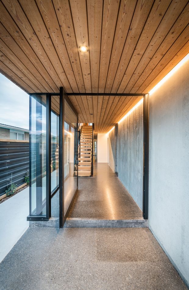A modern covered entry corridor leading to an open-riser staircase. The space features a warm timber-lined ceiling with recessed lighting, polished concrete or terrazzo flooring, exposed concrete walls, black metal framing, extensive floor-to-ceiling glazing, and warm linear wall lighting. An exterior side path and neighbouring building are visible through the glazing.
