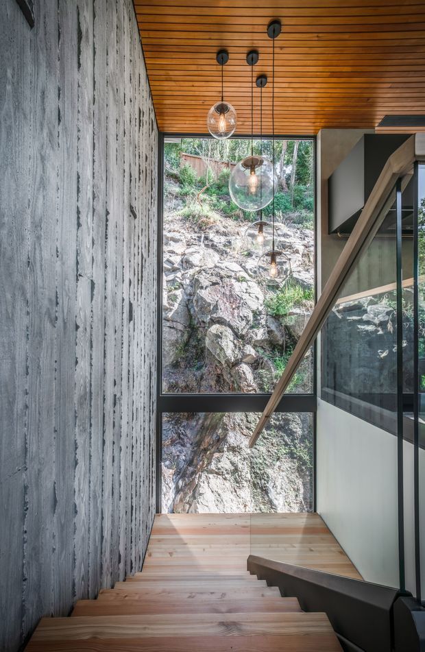 Interior view looking down a contemporary stairway with broad timber treads, a dark handrail and glass balustrade. A heavily textured grey concrete wall contrasts with the warm timber slatted ceiling and stair finishes. Large floor-to-ceiling glazing frames a rocky natural hillside, while multiple suspended globe pendant lights hang through the stair void.
