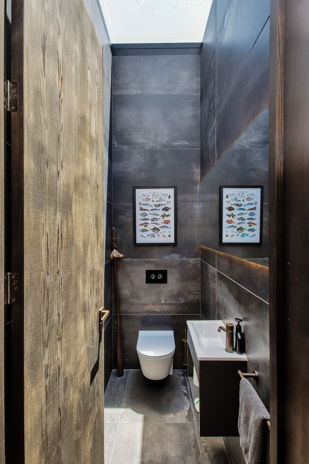 A compact, dark-toned bathroom viewed through an open timber door. The room features large charcoal-grey tiled walls and floor, a wall-hung white toilet, a white rectangular basin on dark built-in cabinetry, a large mirror, black fittings, and framed fish illustrations. A roof skylight brings natural light into the narrow space.