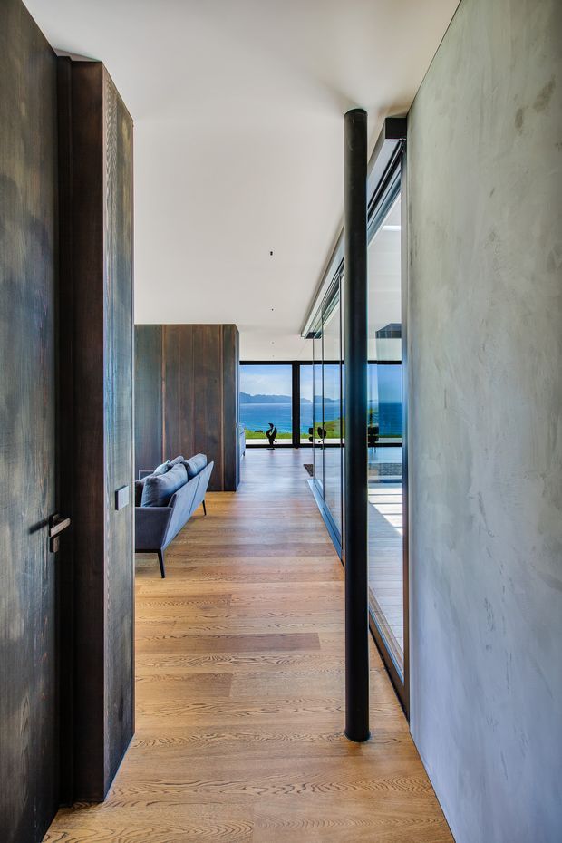 A long, narrow contemporary interior passage leading toward a bright living area and expansive waterfront glazing. The space has wide-plank timber flooring, dark vertical timber doors and wall panels, a smooth light plaster wall, exposed black cylindrical structural columns, and full-height glass overlooking blue water and distant mountains. A sofa is partially visible in the adjoining living room.