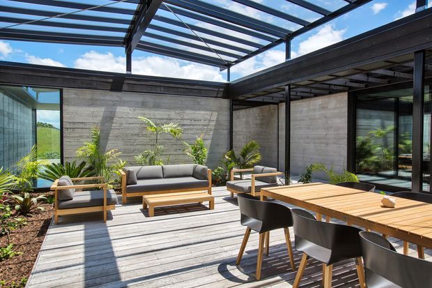 A contemporary outdoor courtyard entertaining area with a timber deck, large timber dining table and black dining chairs, plus a lounge setting with sofas and armchairs. Textured concrete walls, dark metal framing, extensive glazing, and a transparent overhead roof create a sheltered, light-filled outdoor room. Tropical planting lines the perimeter, with open views and blue sky beyond.