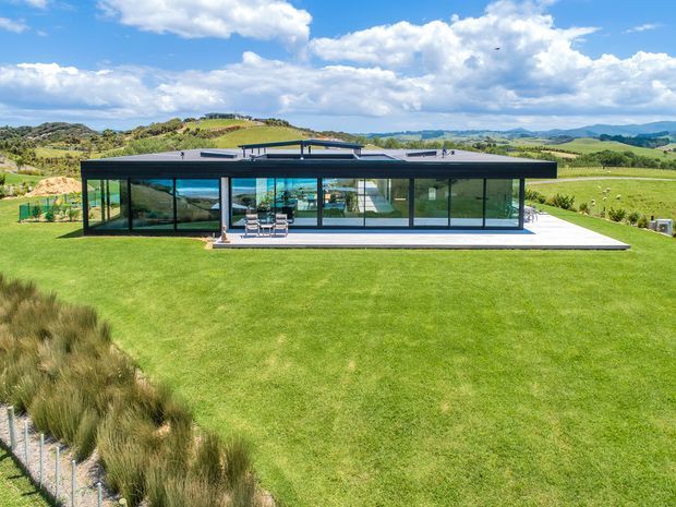 A low-slung contemporary pavilion-style home set in a broad rural landscape. The building has a flat roof, dark slim framing, extensive floor-to-ceiling glazing, and a projecting terrace or paved outdoor area. Rolling green hills and open pasture surround the house.