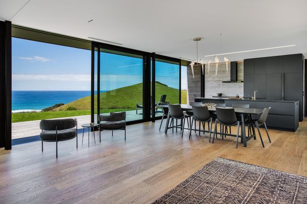 A spacious open-plan contemporary kitchen and dining area with floor-to-ceiling black-framed glazing overlooking a green coastal hillside and ocean. The room features dark minimalist cabinetry, a large island, a dining table with upholstered chairs, timber flooring, pendant lighting, and a neutral patterned rug.