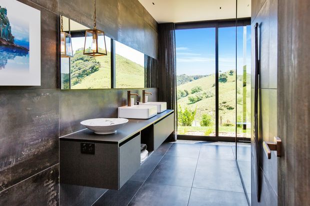 Contemporary bathroom with a floating dark vanity, two white vessel basins, wall-mounted fixtures, large-format grey floor and wall finishes, and full-height glazing overlooking a green hilly landscape. Black-framed glass doors, pendant lights, and a large mirror create a refined, dramatic interior.
