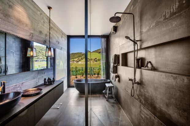 Industrial-style bathroom with a freestanding dark bathtub positioned beside full-height glazing overlooking a green hillside. The room features concrete-look walls, large grey floor tiles, a long dark vanity with vessel basins, black framed shower glazing, exposed black plumbing fixtures, and warm pendant lighting.