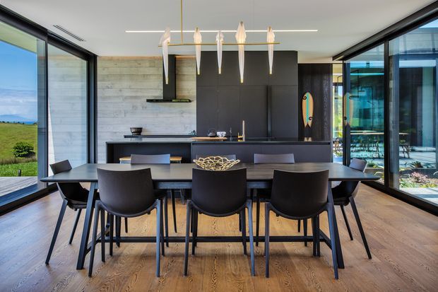 A contemporary open-plan dining and kitchen area with a long dark dining table surrounded by black chairs. Floor-to-ceiling glazing on both sides provides expansive views, while a compact black kitchen with a concrete-look backsplash, integrated appliances and pendant lighting forms the backdrop. Warm timber flooring contrasts with the dark cabinetry and furniture.