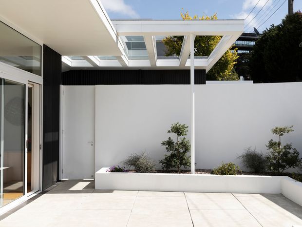 A bright contemporary courtyard or terrace enclosed by white rendered walls, with large pale paving slabs, raised integrated garden beds, and sparse ornamental planting. A white pergola with translucent roof panels provides overhead shelter, while dark vertical cladding contrasts with extensive glazed sliding doors at the left. The space feels private, clean, and minimalist, with glimpses of surrounding urban buildings beyond the walls.