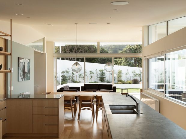 A bright, open-plan contemporary kitchen and dining area with a long concrete island, integrated sink, warm timber cabinetry, and a timber dining table with chairs. Floor-to-ceiling glazing opens to a landscaped outdoor area, while a lounge seating area is visible beyond. Pendant lights and recessed ceiling lights provide a clean, understated fit-out.