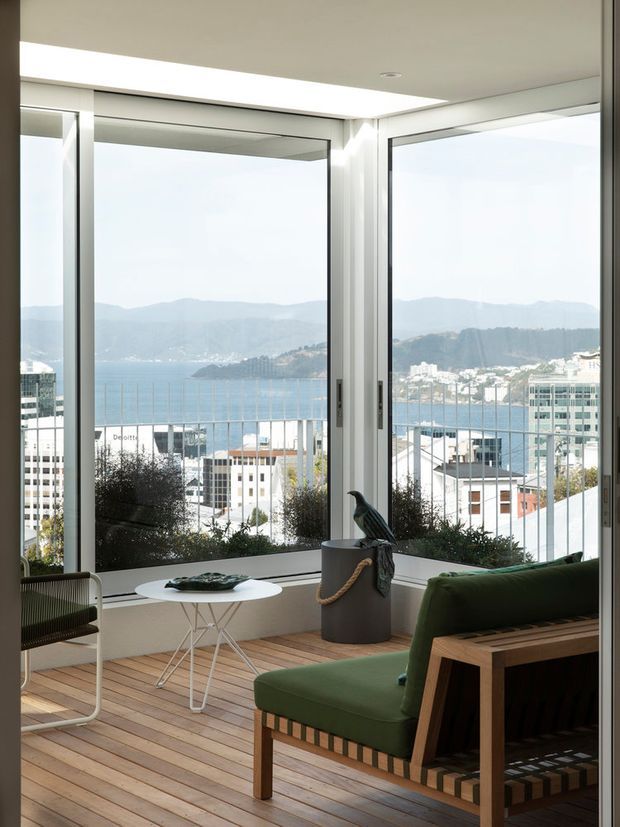 A bright, compact sunroom or enclosed balcony with expansive corner glazing overlooking a bay, hills, and dense urban buildings. The space has a warm timber plank floor, white walls and window frames, a green upholstered timber-framed lounge chair, a woven chair, a small white side table, and a sculptural bird object. The clean-lined glazing and restrained furnishings create a calm mid-century-inspired coastal atmosphere.