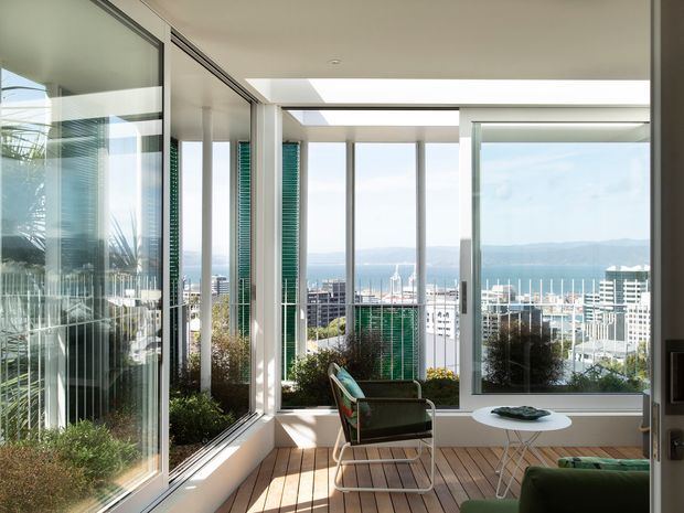 Bright enclosed sunroom or glazed balcony overlooking a waterfront city and harbour. The space features extensive floor-to-ceiling glazing, white structural framing, timber decking, a compact lounge chair and low table, and strong indoor-outdoor visual connection. The image has a clean contemporary architectural character with abundant natural light.