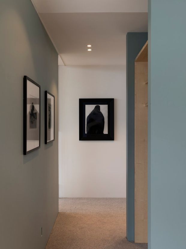 A narrow, softly lit residential hallway with muted grey and blue painted walls, a pale ceiling with recessed downlights, beige carpet flooring, framed artwork along the left wall, and a light timber built-in or doorway feature on the right. The corridor leads toward a white end wall and has a calm, understated contemporary feel.