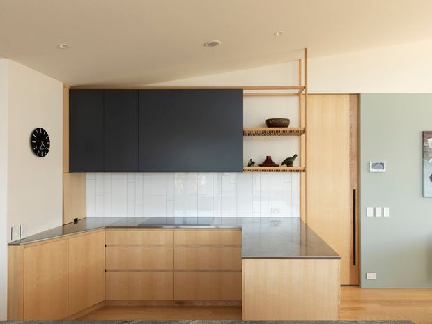 A compact contemporary kitchen with light timber cabinetry, a dark charcoal overhead cabinet block, white vertical tile splashback, and a dark stone island benchtop. Open timber shelving displays a few decorative objects, while a timber door and pale green feature wall are visible to the right. The kitchen has a clean, warm, streamlined appearance with integrated appliances and minimal detailing.
